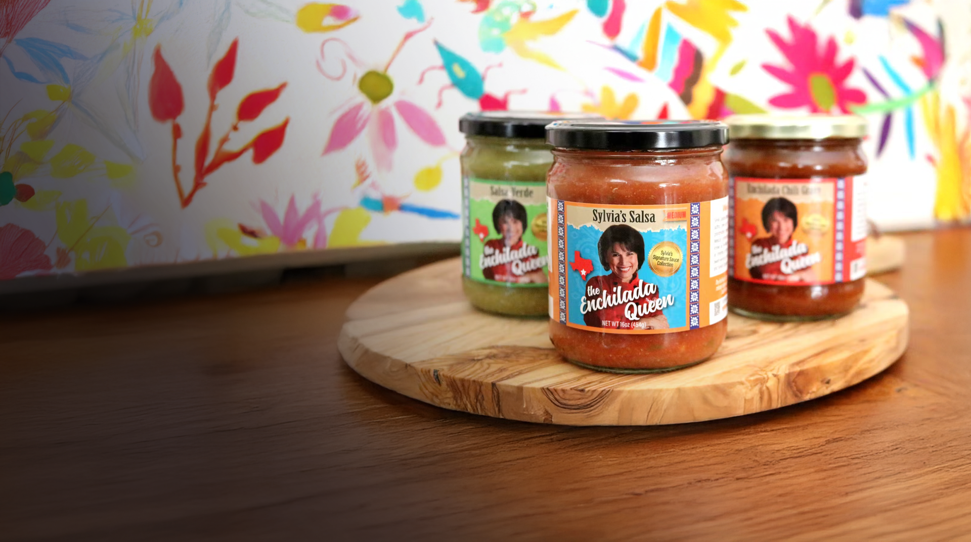 Three jars of salsa on a wooden board with a colorful floral-patterned appliance in the background.